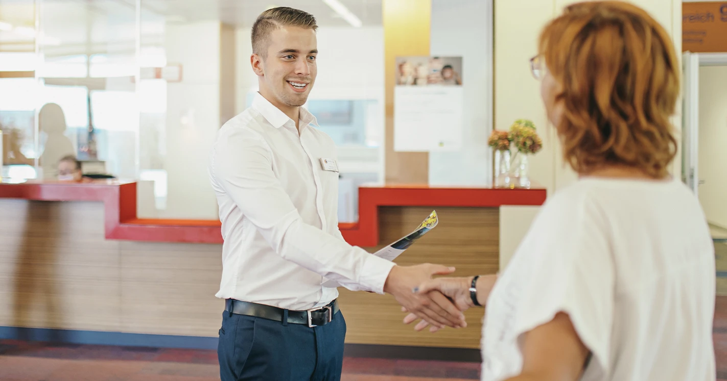 Ein Mitarbeiter vom Empfang begrüßt freundlich eine Rehabilitandin.
